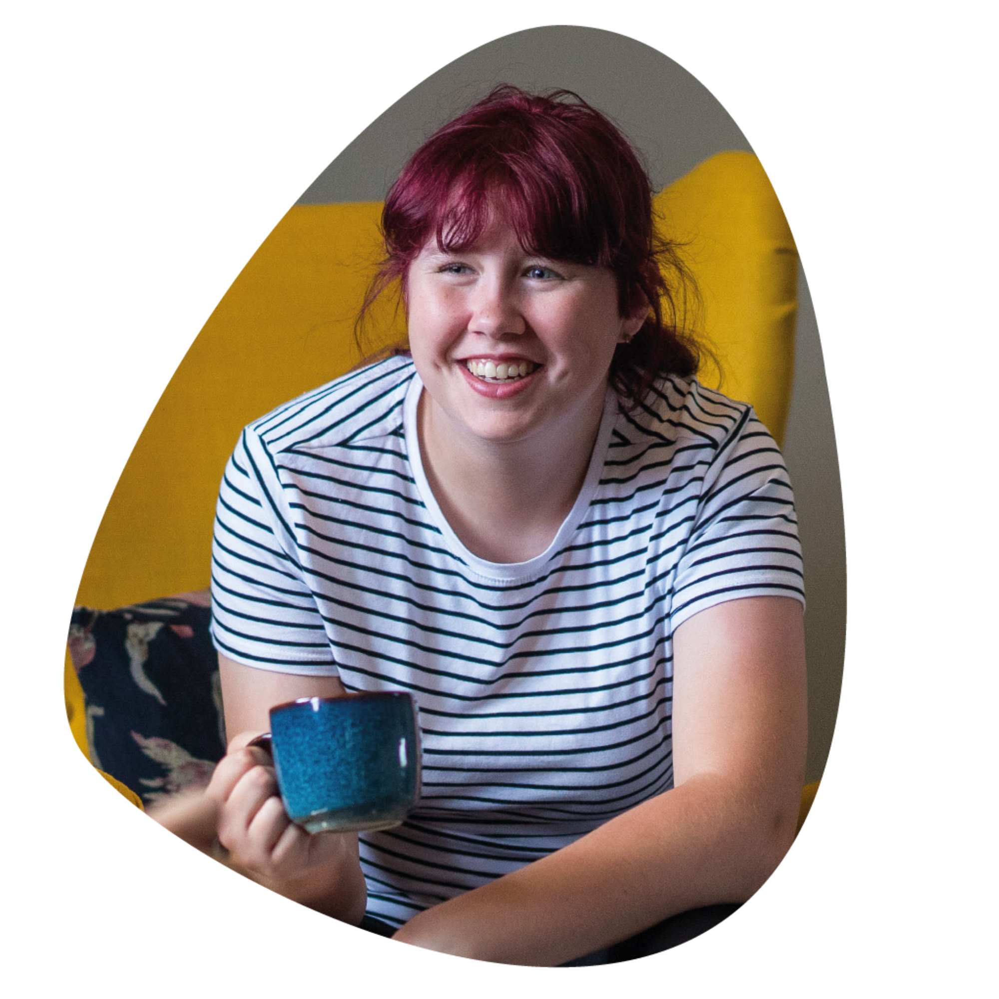 A smiling Creating Tomorrow College learner sitting indoors holding a mug, wearing a striped T-shirt, and looking forward toward someone off camera in a relaxed, friendly setting,