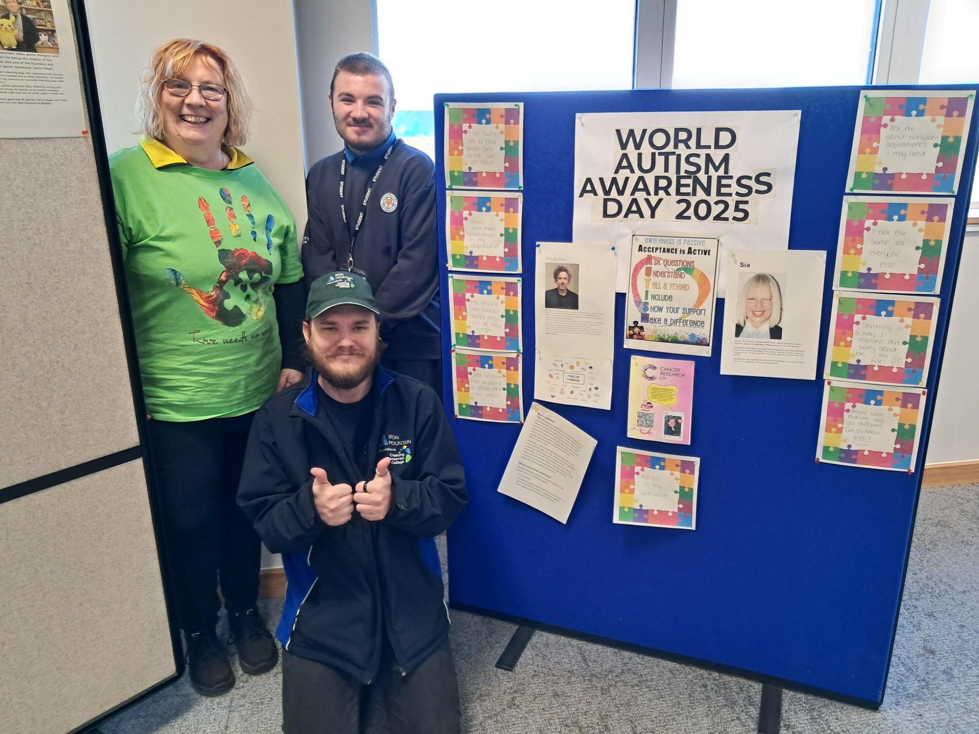 Two Creating Tomorrow College learners and Alice from Iron Mountain standing beside a blue display board titled World Autism Day 2025.