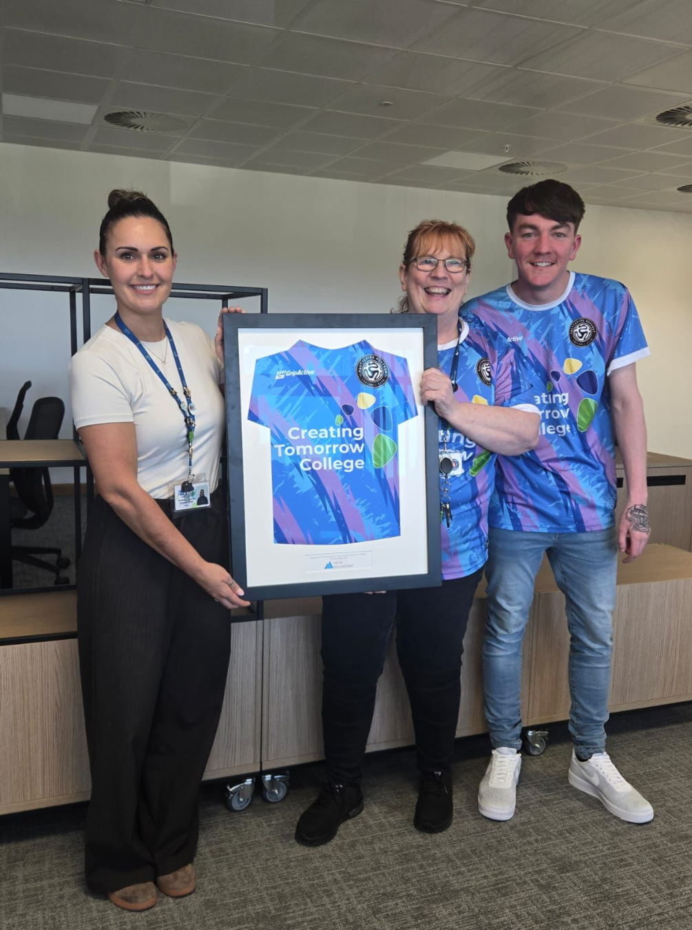 Creating Tomorrow College Kettering Campus Leader, Representative from Iron Mountain and Representative from Kettering Rovers FC all standing together, smiling and holding a framed shirt
