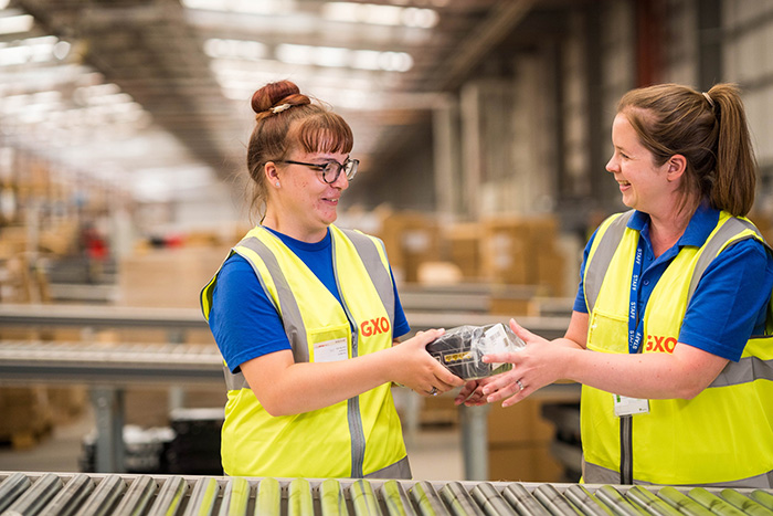 A photo of a student and job coach working in high viz jackets in a factory