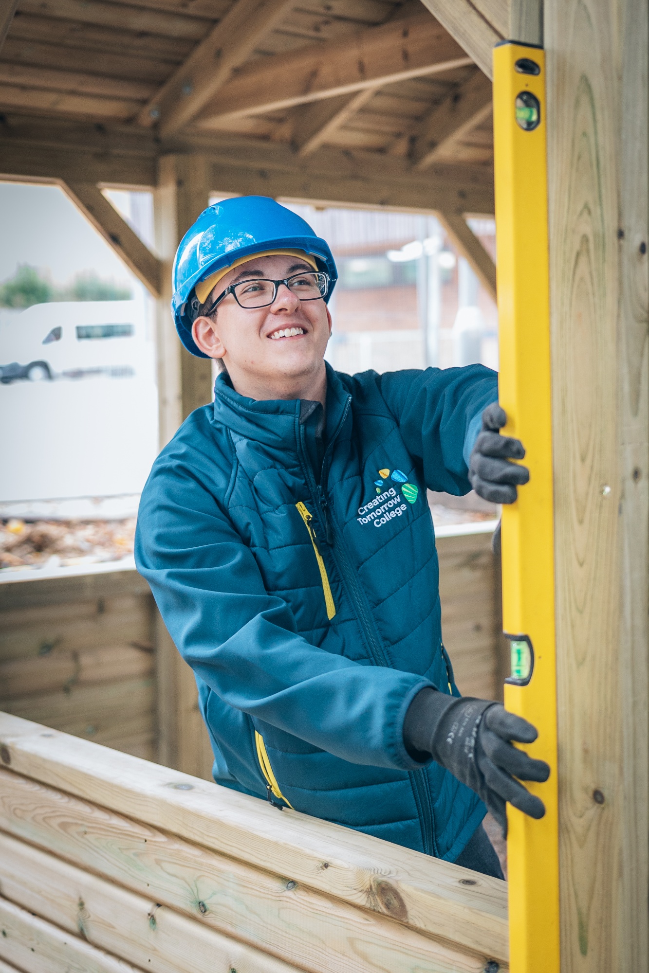 Student wearing a blue hard hat and safety gloves uses spirit level to check a wooden structure while smiling, at Creating Tomorrow College