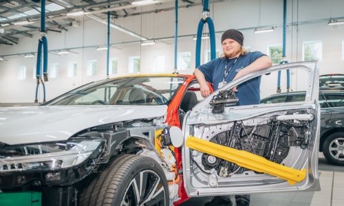 A learner wearing a beanie hat and Creating Tomorrow College uniform standing beside a partially disassembled car in a bright automotive workshop