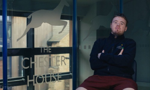 Tyrone sitting outside the Wellingborough Campus, next to a gall wall with the Chester House logo